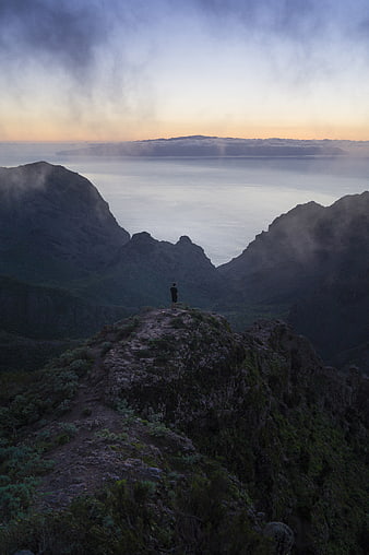 Man standing on mountain peak overlooking ocean at dusk