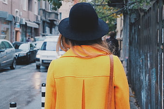 Woman in Yellow Coat and Black Hat Walking Down City Street