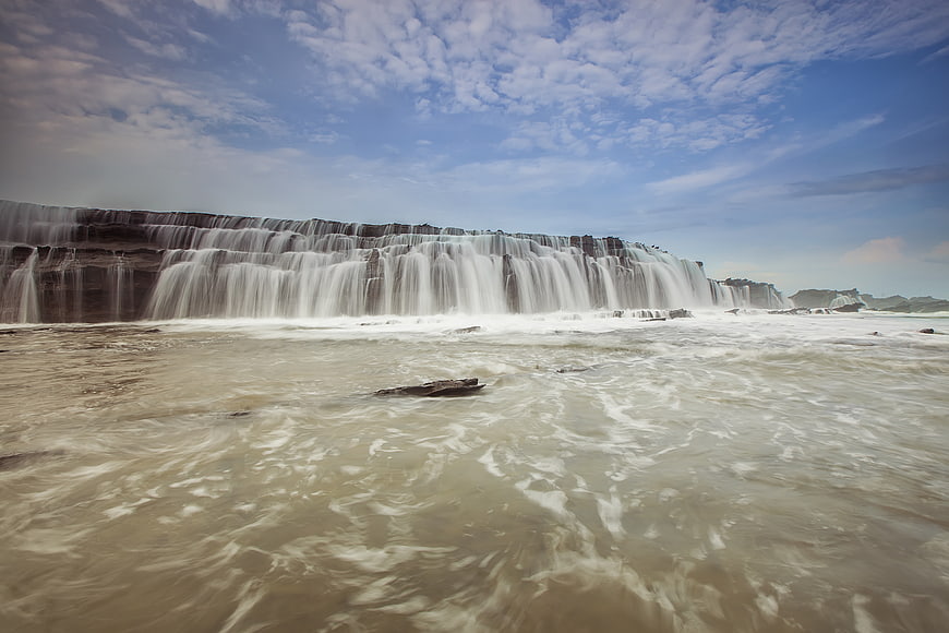 Wide waterfall cascading into a river under a blue sky