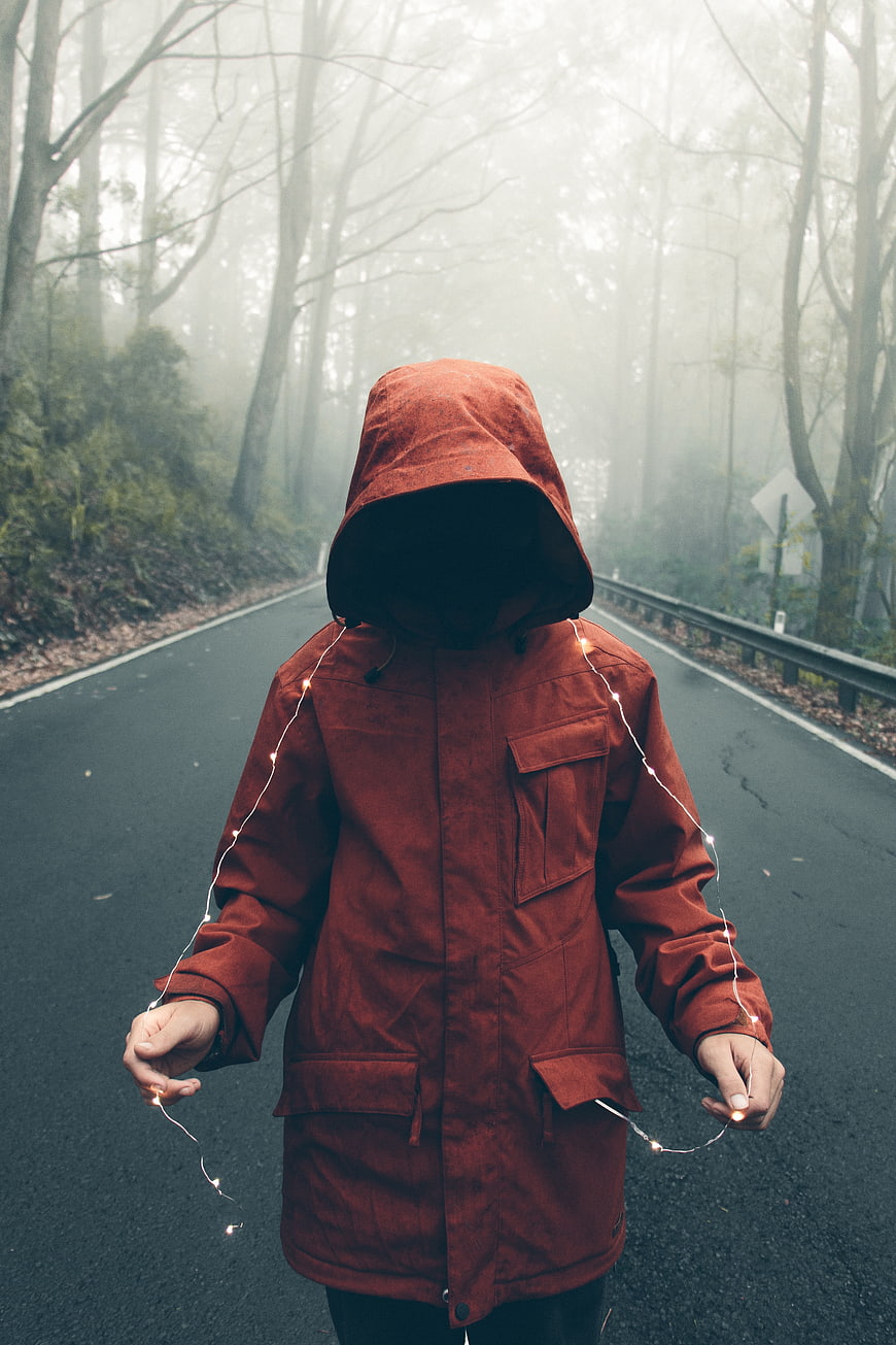 Mysterious hooded figure with fairy lights on foggy forest road