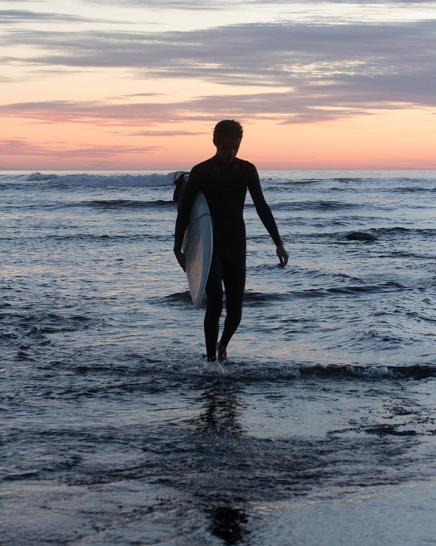 Surfer walking out of ocean at sunset with surfboard