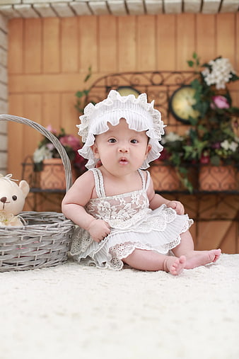 Adorable Baby Girl in White Lace Dress and Bonnet Sitting on Rug