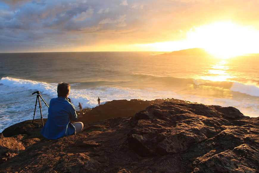 Photographer on Rocky Cliff Capturing Golden Sunset Over Ocean