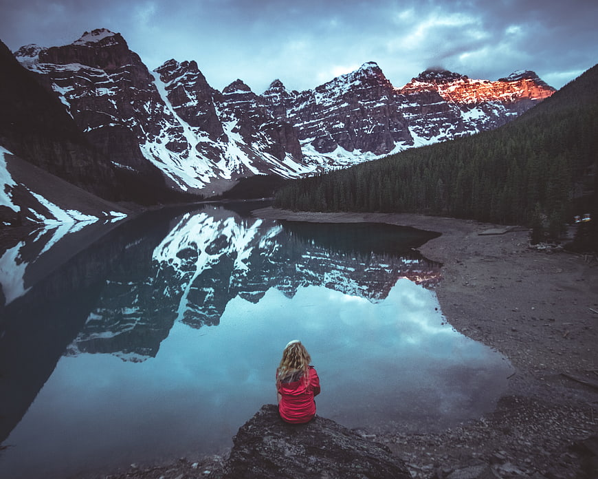 Woman in Red Jacket Sitting by Mountain Lake Reflection