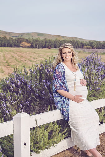 Pregnant woman in white dress leaning on white fence in lavender field