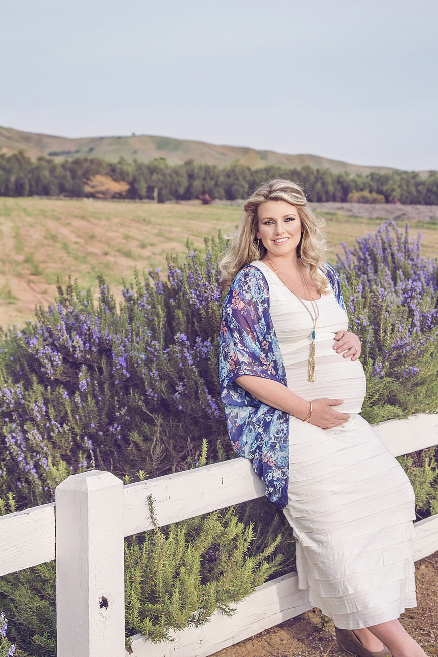 Pregnant woman in white dress leaning on white fence in lavender field