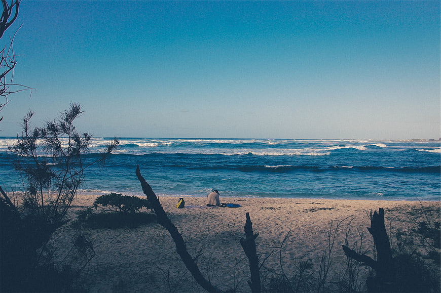 Lone figure sitting on sandy beach with blue ocean waves
