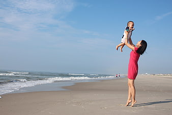 Mother lifting baby on sunny beach with ocean waves