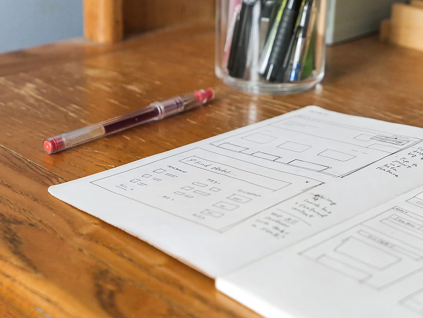 Hand-drawn UI wireframes on notebook with red pen on wooden desk