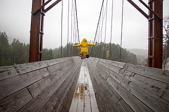 Woman in Yellow Raincoat Jumping on Wooden Suspension Bridge