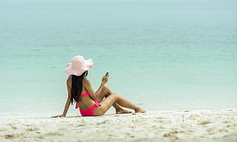 Woman in pink bikini and hat using smartphone on tropical beach