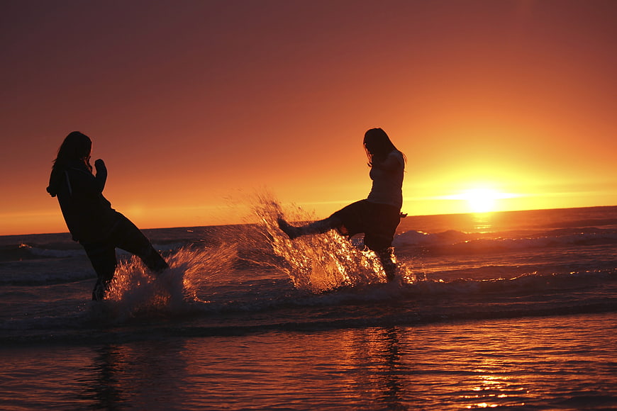 Two women splashing water on beach at sunset