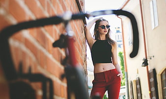Stylish woman in red pants leaning against brick wall with bicycle