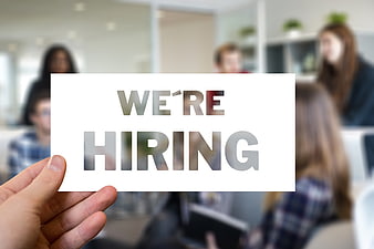 Hand holding We're Hiring sign in blurred office background