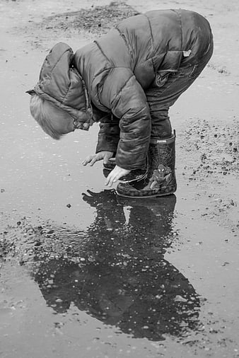 Child in rain boots touching puddle reflection black and white