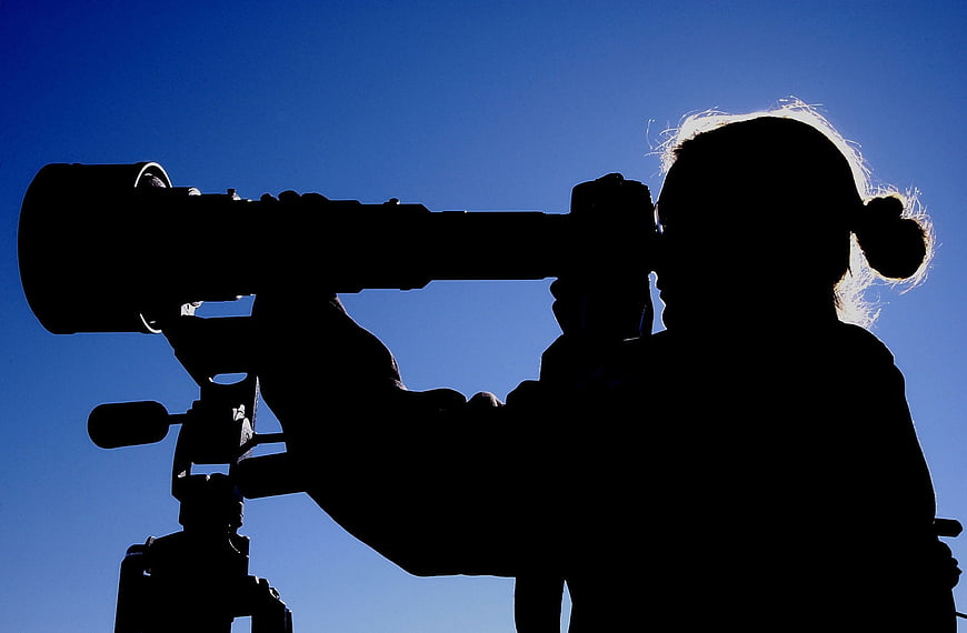 Silhouette of woman using large camera lens against blue sky