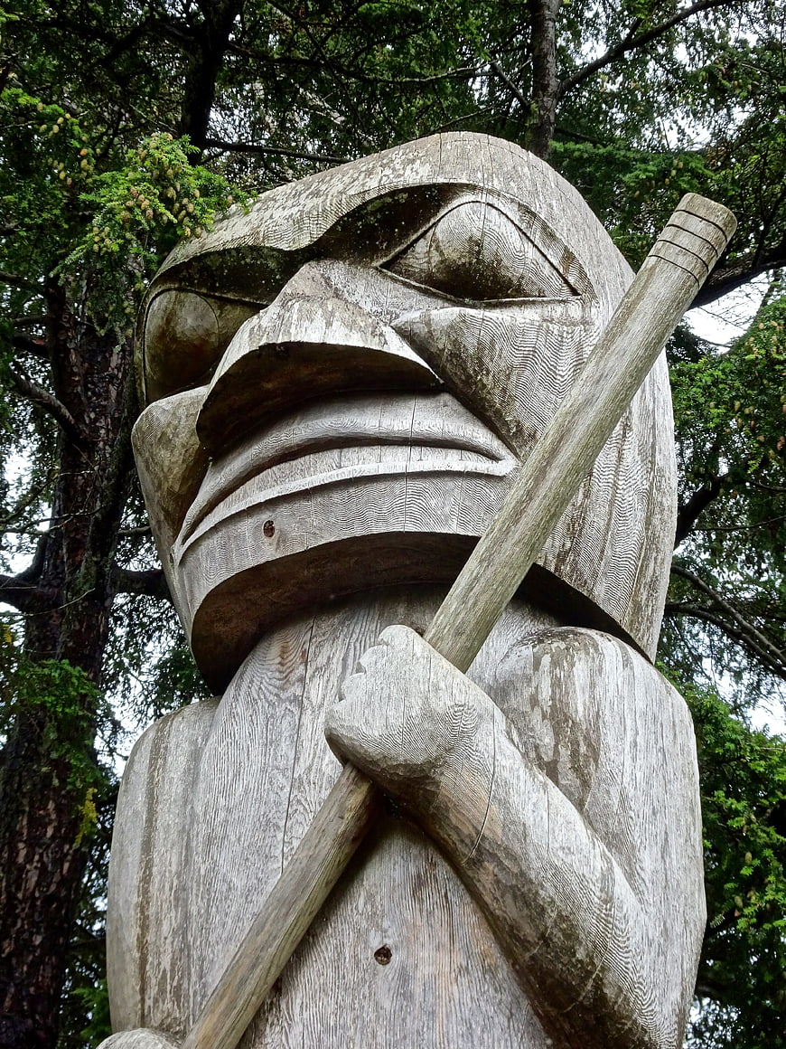 Weathered wooden totem pole figure holding a staff in a forest