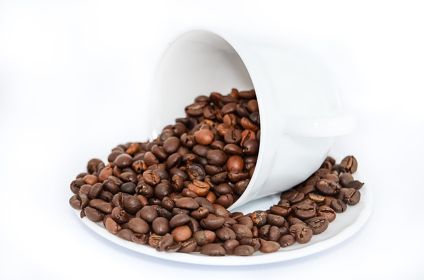 Roasted coffee beans spilling from white ceramic cup on saucer