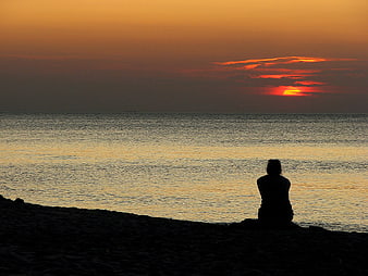 Silhouette of person sitting on beach watching sunset over ocean