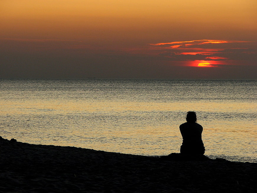 Silhouette of person sitting on beach watching sunset over ocean