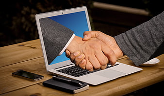 Handshake emerging from laptop screen on wooden table
