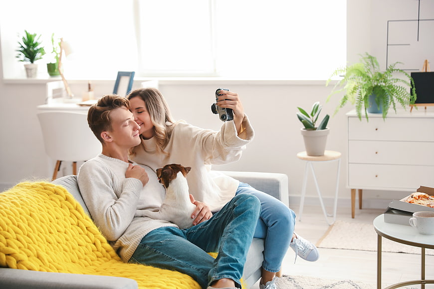 Happy couple taking selfie with vintage camera and dog on sofa