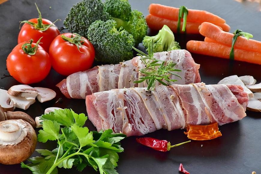 Raw pork tenderloin tied with bacon and fresh vegetables on dark slate