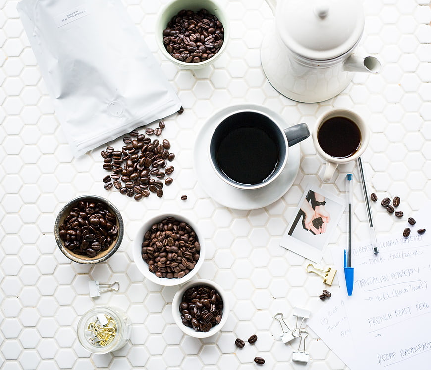 Flat lay of coffee beans, cups, and office supplies on white tiles