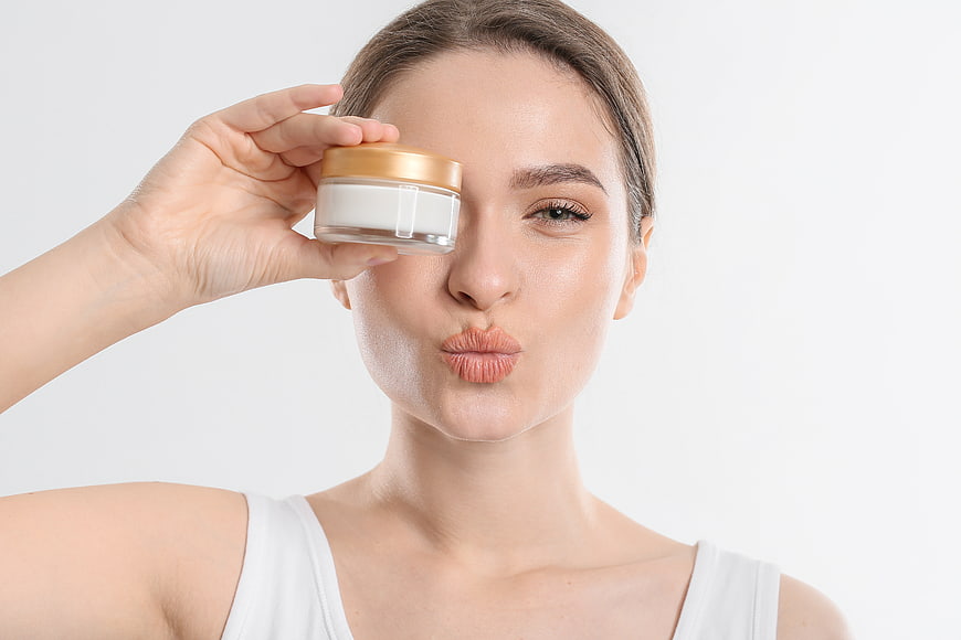 Playful woman holding skincare cream jar over eye