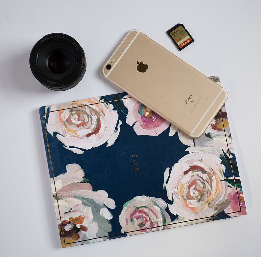 Flat lay of gold iPhone, camera lens, and floral planner on white background