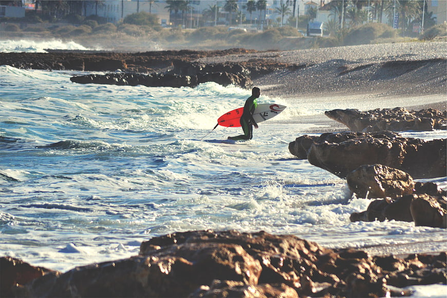 Surfer with Red Board Walking Through Rocky Ocean Waves