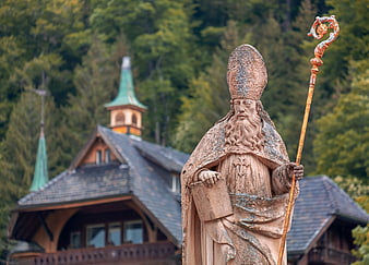 Stone statue of Saint Blaise with staff in front of traditional Black Forest church