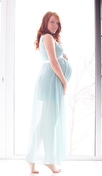 Pregnant woman in flowing mint dress standing by bright window