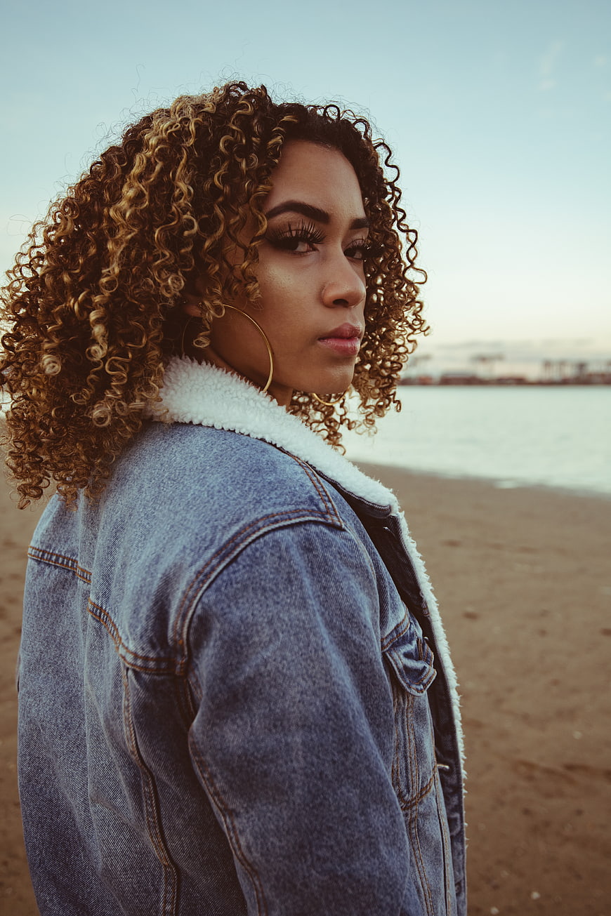 Stylish Woman with Curly Hair on Beach at Sunset