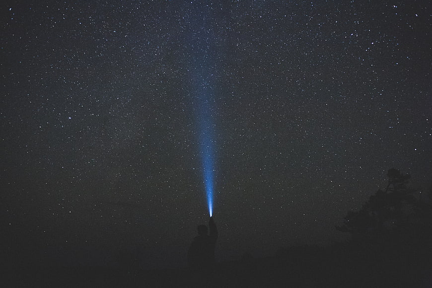 Silhouette of Person Holding Flashlight Beam into Starry Night Sky