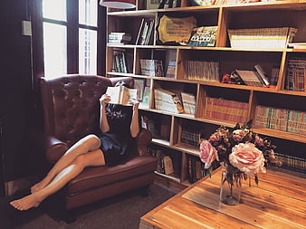 Woman reading book in leather armchair near bookshelves