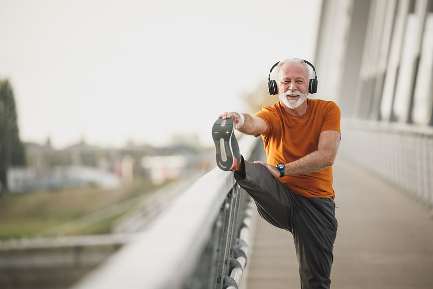 Senior man stretching leg on bridge railing with headphones