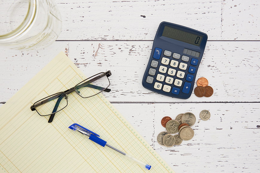 Flat lay of calculator, coins, glasses, and notepad on white wood