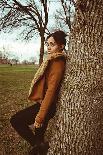 Young woman in brown jacket leaning against tree in park