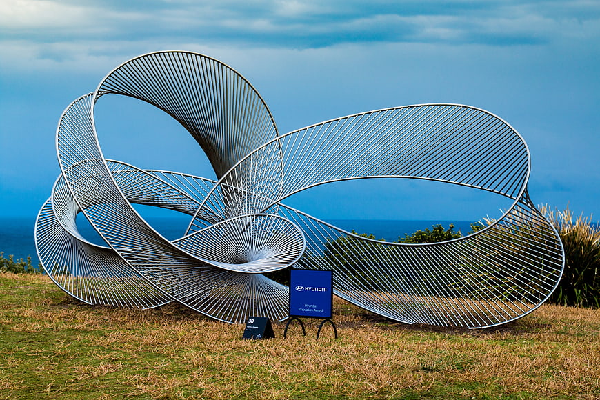 Modern metal spiral sculpture on grassy hill overlooking ocean