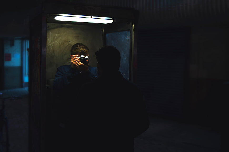Man Taking Mirror Selfie with Camera in Dark Phone Booth