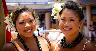 Two smiling Polynesian women with white lilies and black beaded necklaces