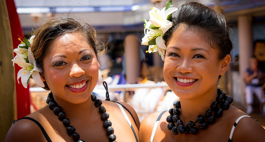 Two smiling Polynesian women with white lilies and black beaded necklaces