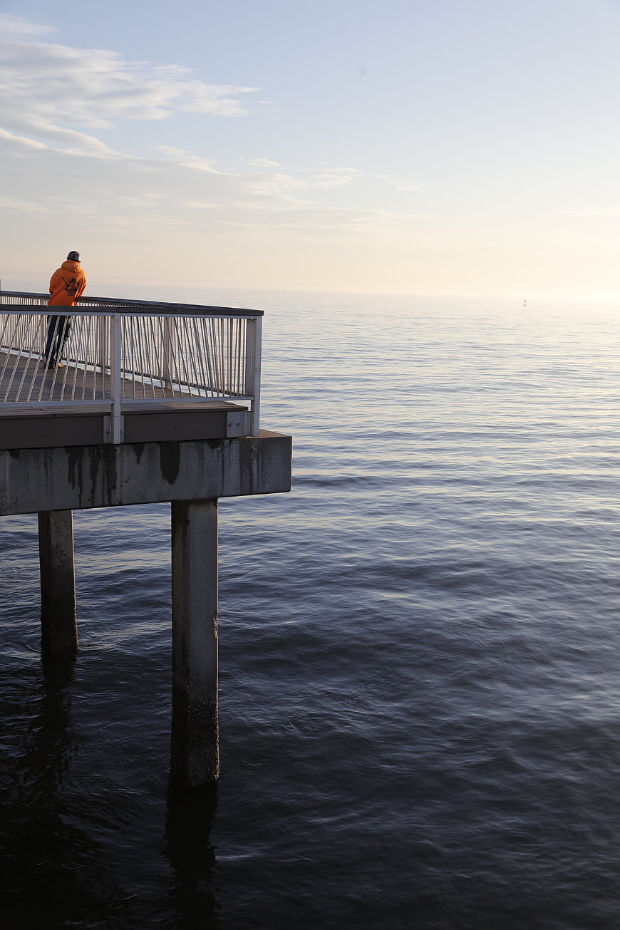 Man in Orange Jacket Standing on Pier Overlooking Calm Sea
