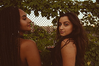 Two young women posing outdoors near a vine-covered fence