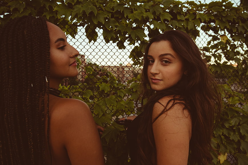 Two young women posing outdoors near a vine-covered fence