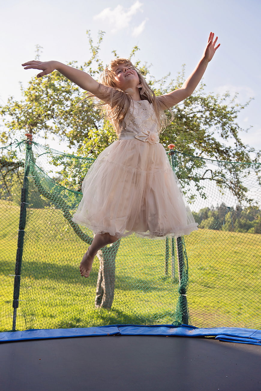 Young girl in beige dress jumping on trampoline outdoors