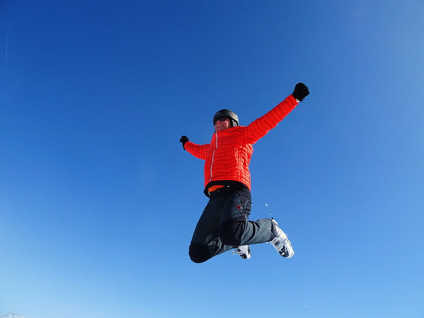 Man in Orange Jacket Jumping Against Blue Sky