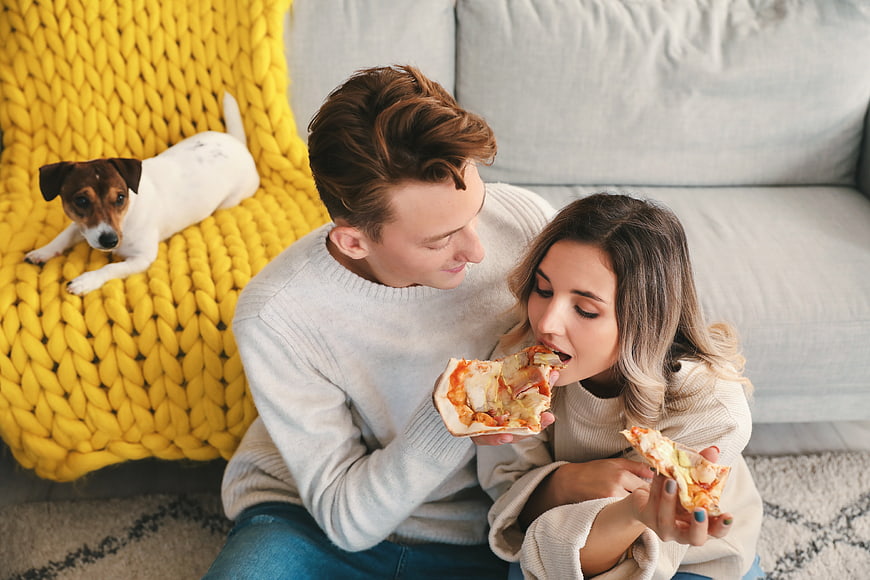 Young couple eating pizza on floor with dog