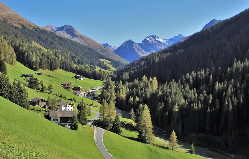 Alpine Village Landscape With Winding Road And Snow-Capped Peaks